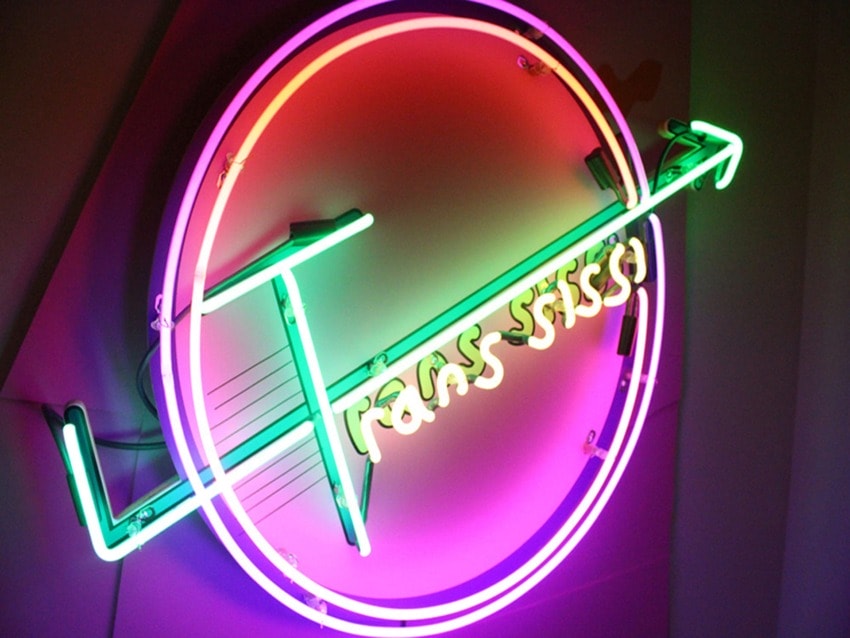 Colorful neon light art with circular and arrow patterns, featuring the text "Transmissions" in vibrant pink and green hues.