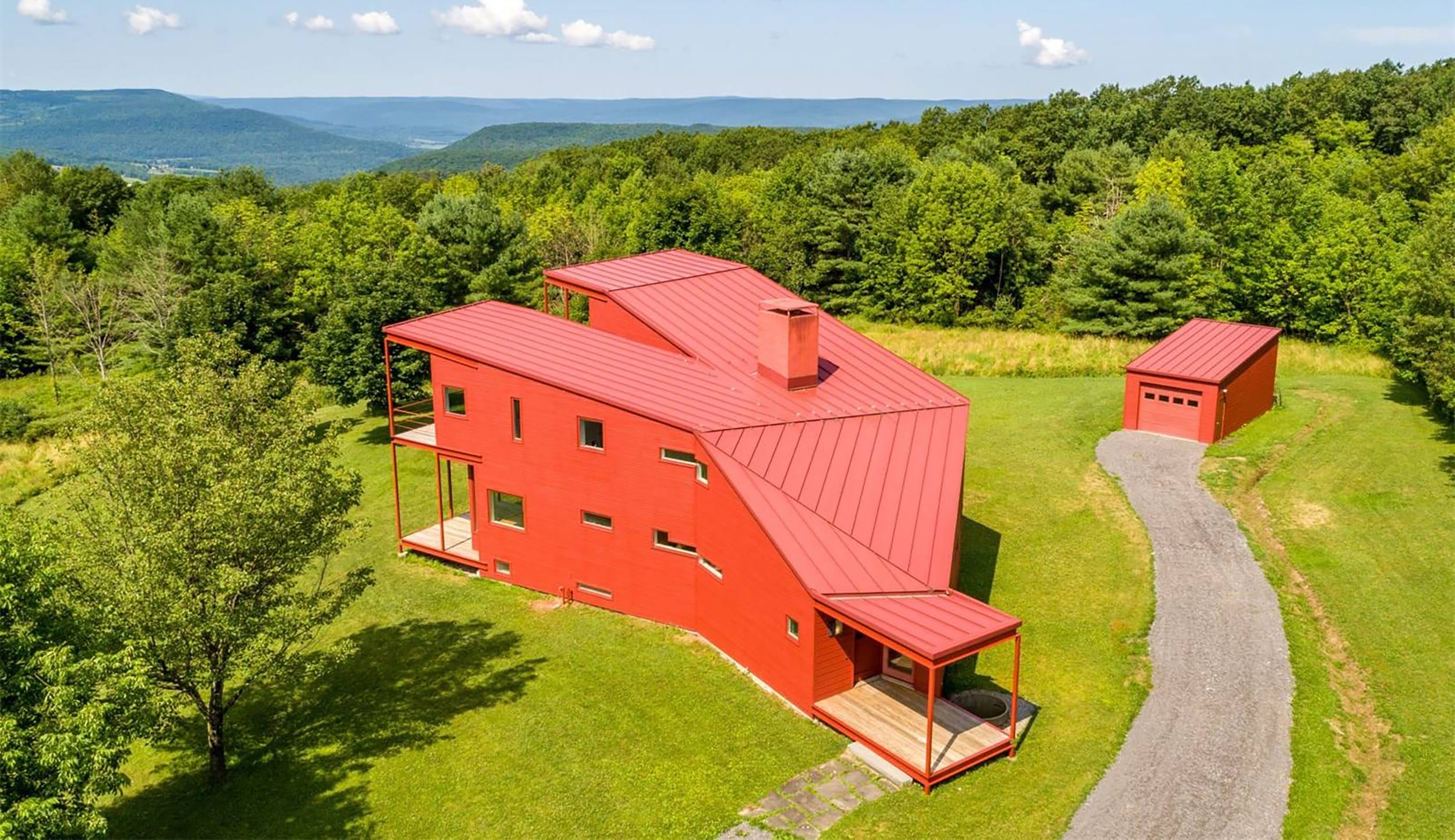 Aerial view of a modern red house with a metal roof in a lush green landscape with hills in the background.