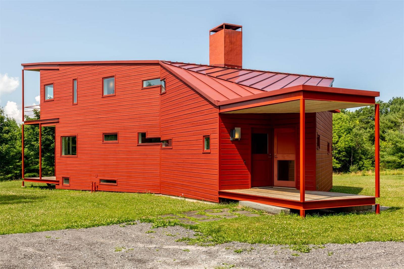 Red modern house with a unique angular design, featuring a wraparound porch and surrounded by a grassy lawn.
