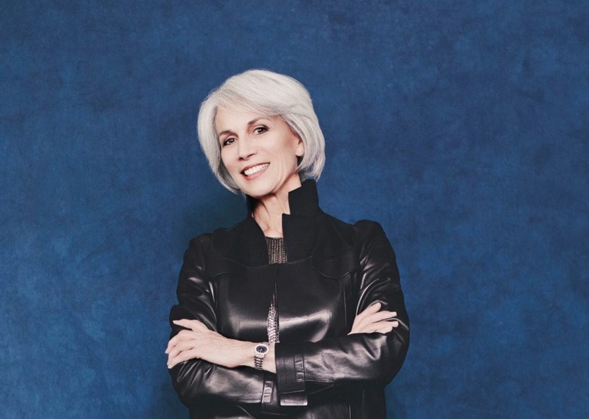 Smiling woman with short white hair in a black jacket stands against a solid blue background.