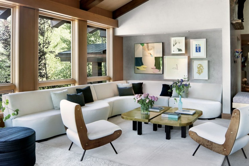 Modern living room with white sectional sofa, wooden accent chairs, green tables, large windows, and art on the wall.