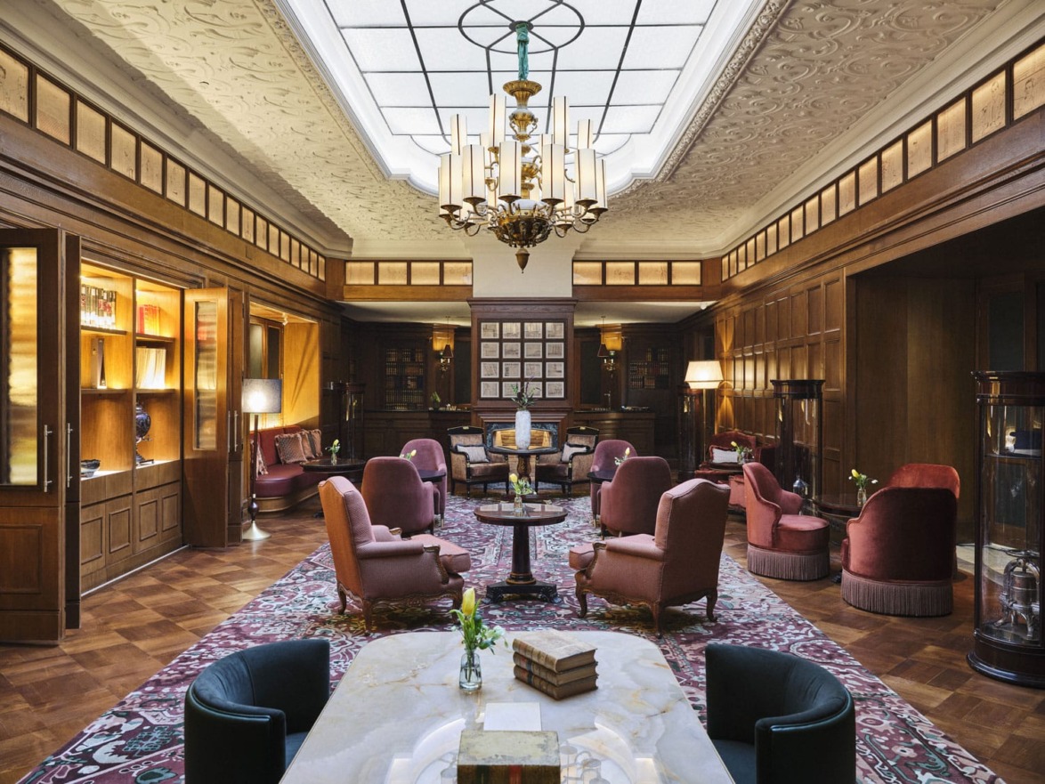 Elegant hotel lounge with vintage decor, chandelier, plush chairs, wooden bookshelves, and a patterned rug under soft lighting.