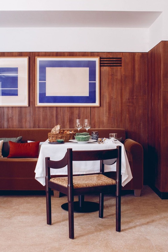 Cozy dining area with a wooden chair and table set for two, featuring modern art on wood-paneled walls.