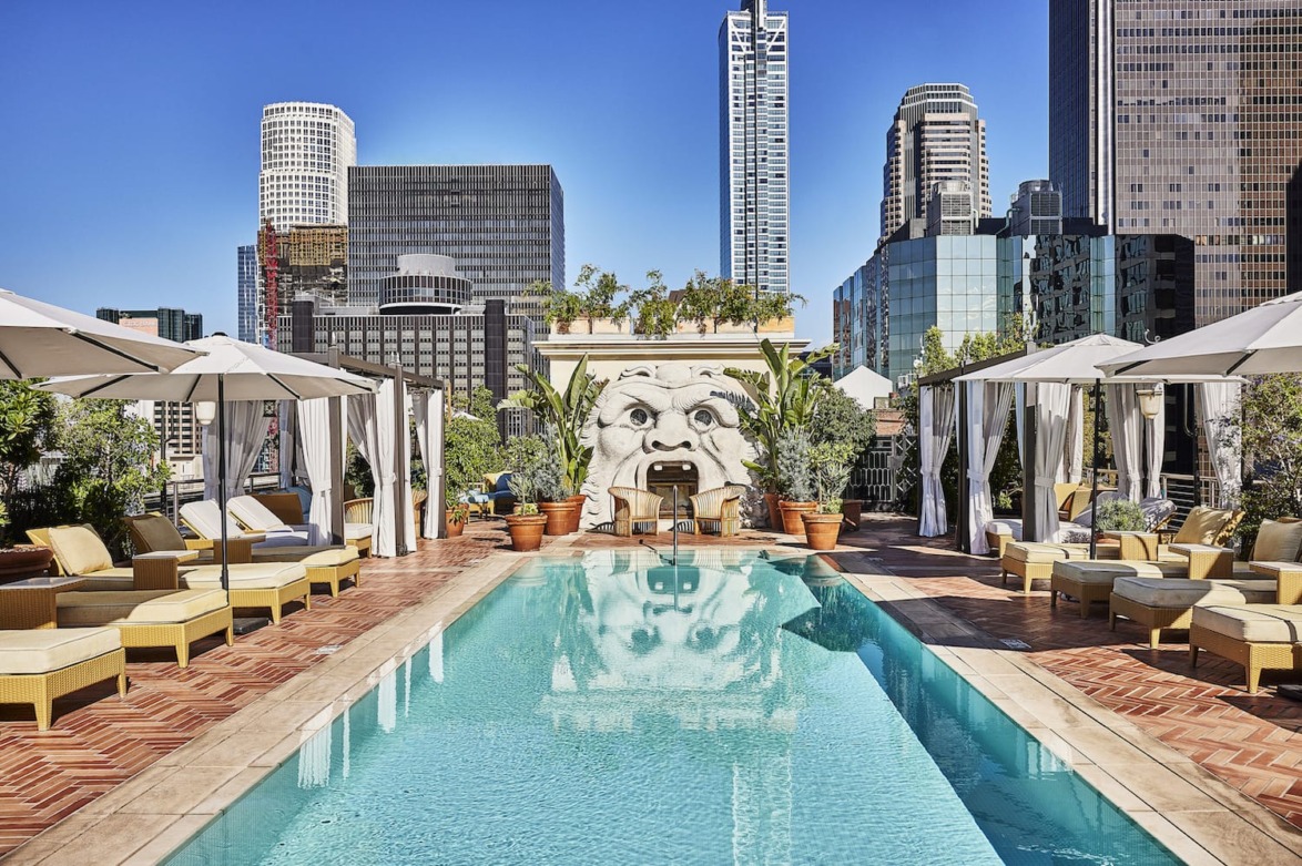 Rooftop pool with lounge chairs and cabanas, city skyscrapers in the background under a clear blue sky.