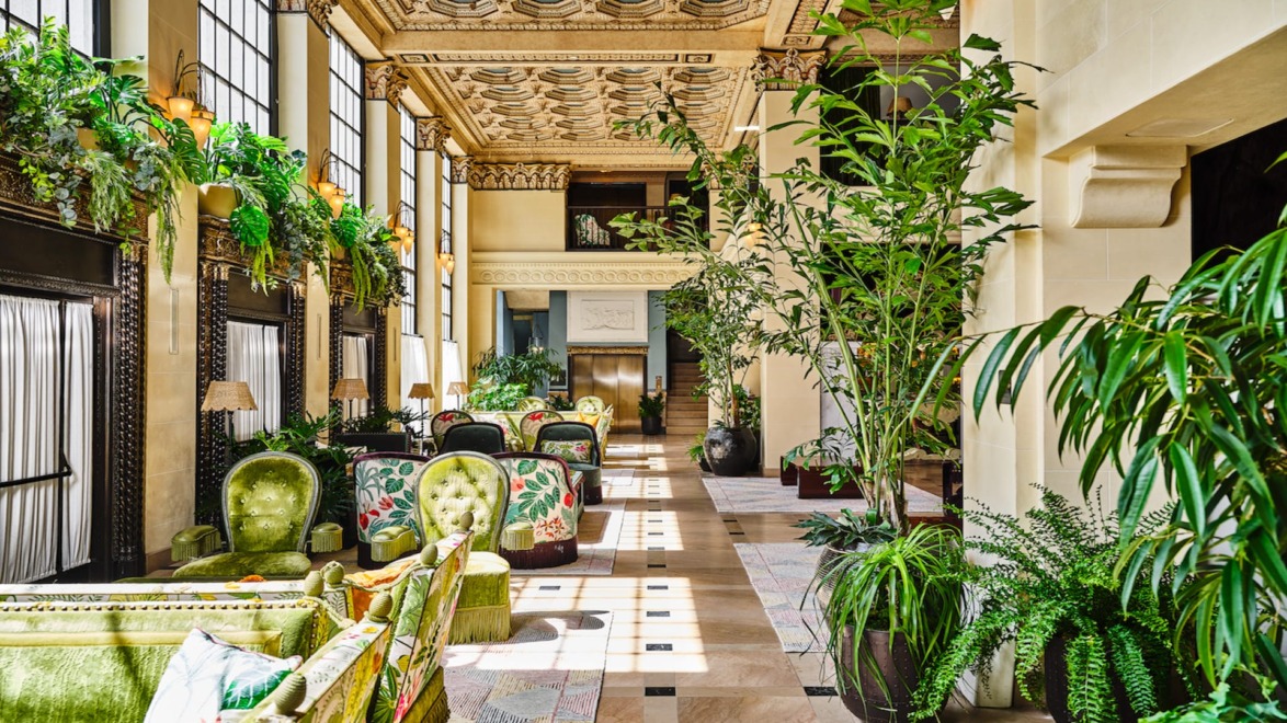 Luxurious hotel lobby with ornate ceiling, lush green plants, and elegant seating areas bathed in natural light.