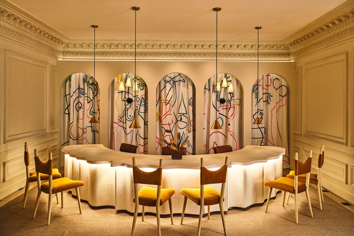 Elegant dining room with modern art backdrop, curved illuminated table, and six wooden chairs with yellow cushions.