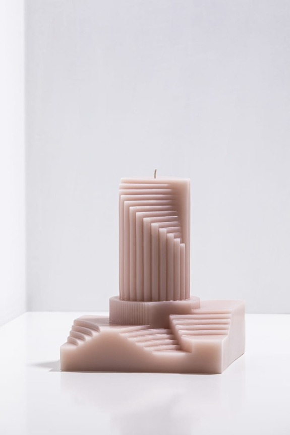 Beige sculptural candle with geometric stair-step design placed on a white surface against a plain background.