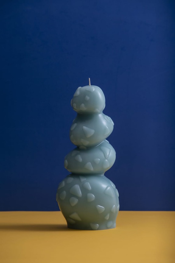 Unique blue candle with abstract shapes stacked vertically, set against a blue background on a yellow surface.