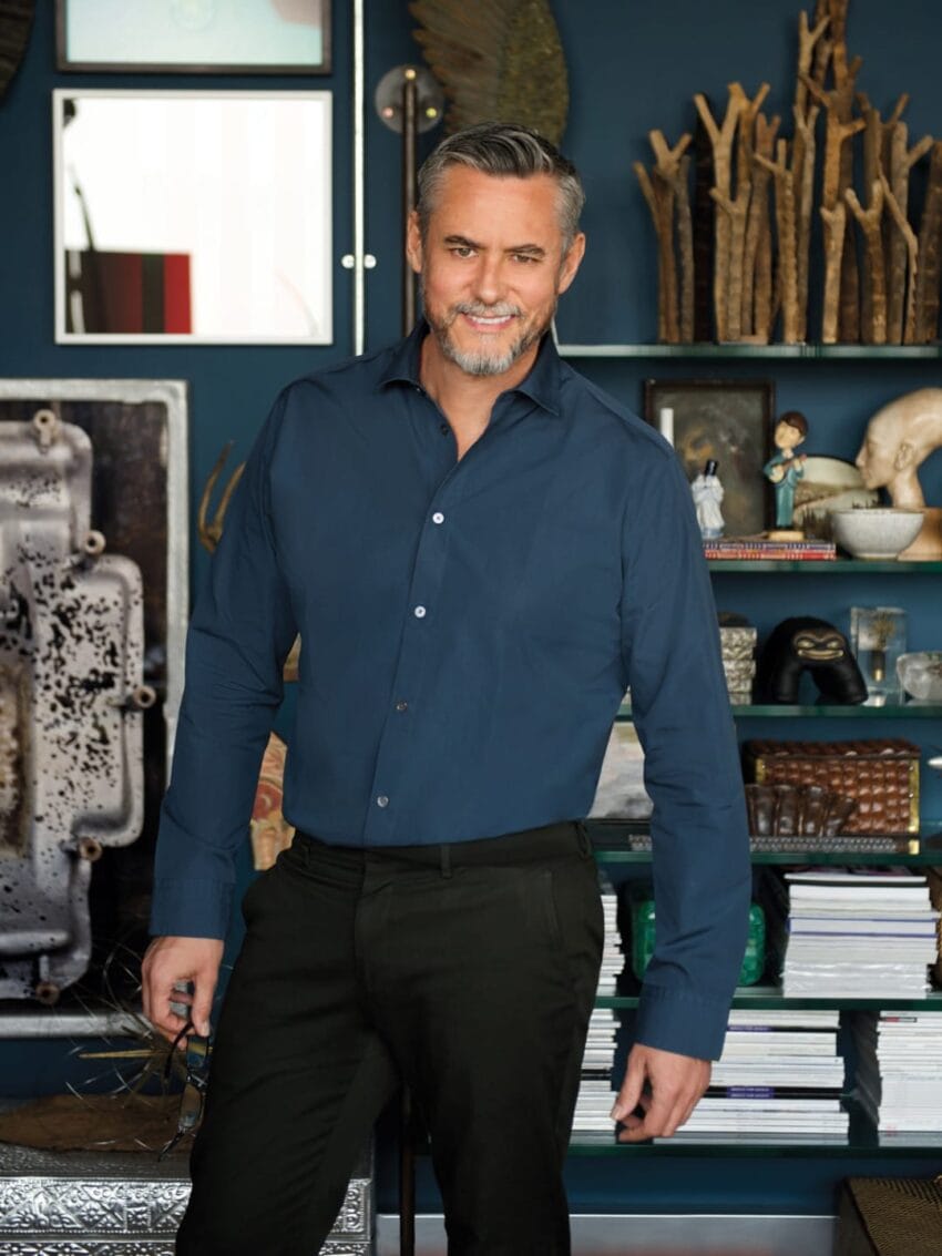 Man with gray hair and beard standing in a room with bookshelves and eclectic decor, wearing a dark shirt and pants.