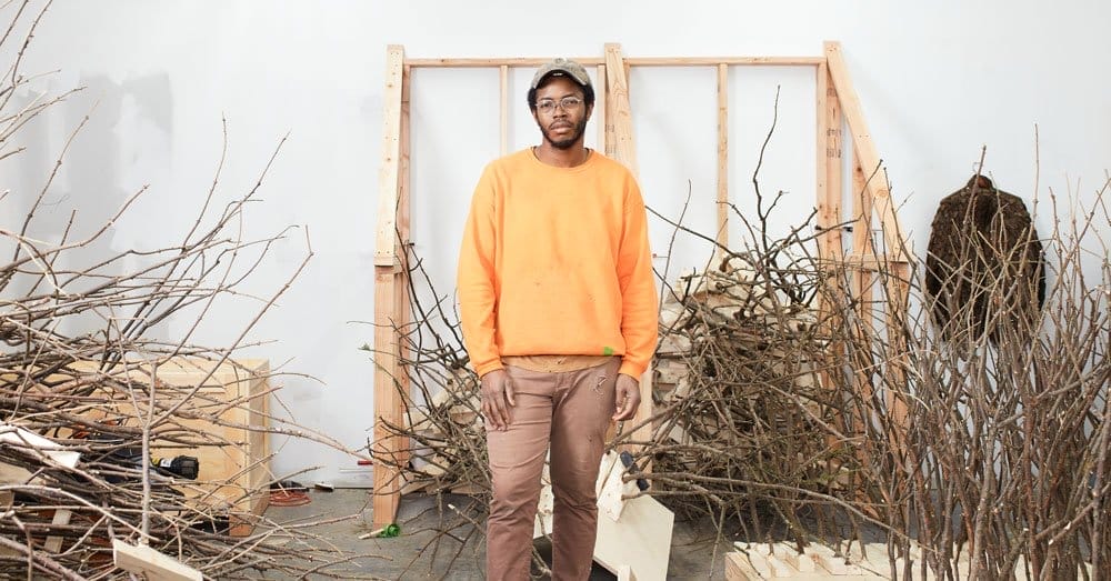Person standing in a workshop surrounded by stacks of branches, wearing an orange sweater and cap.