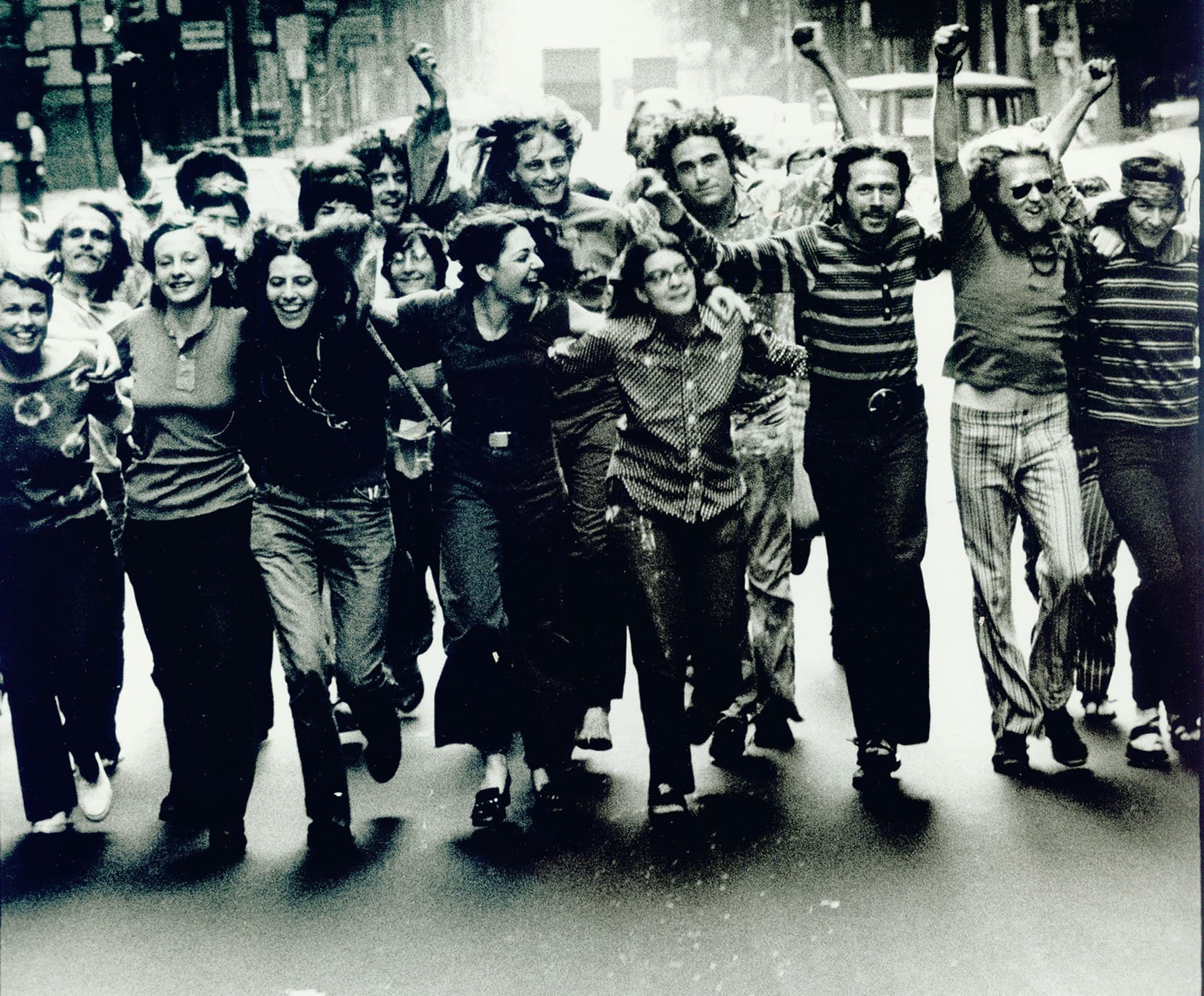 Group of people marching arm in arm, raising fists in a lively city street, showcasing unity and determination.