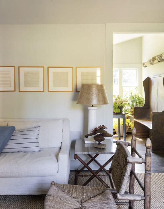 Cozy living room with white sofa, framed art, rustic chairs, and a lamp on a side table near a sunlit doorway.