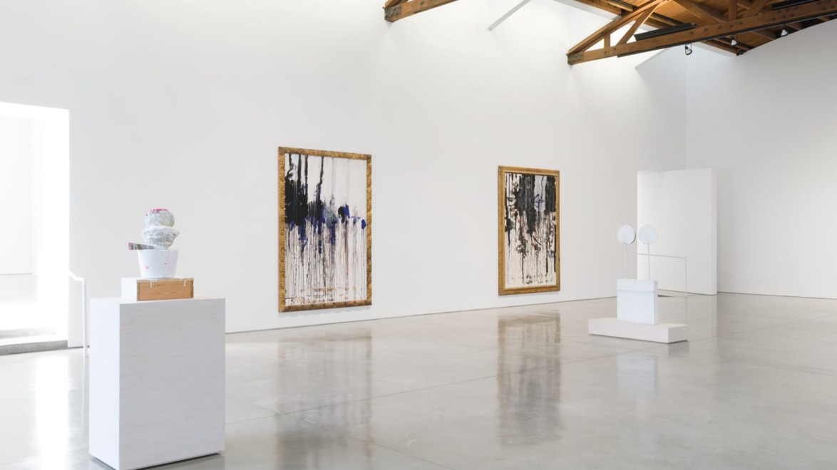 Art gallery interior with large abstract paintings on white walls, ceramic sculptures on pedestals, and natural light.