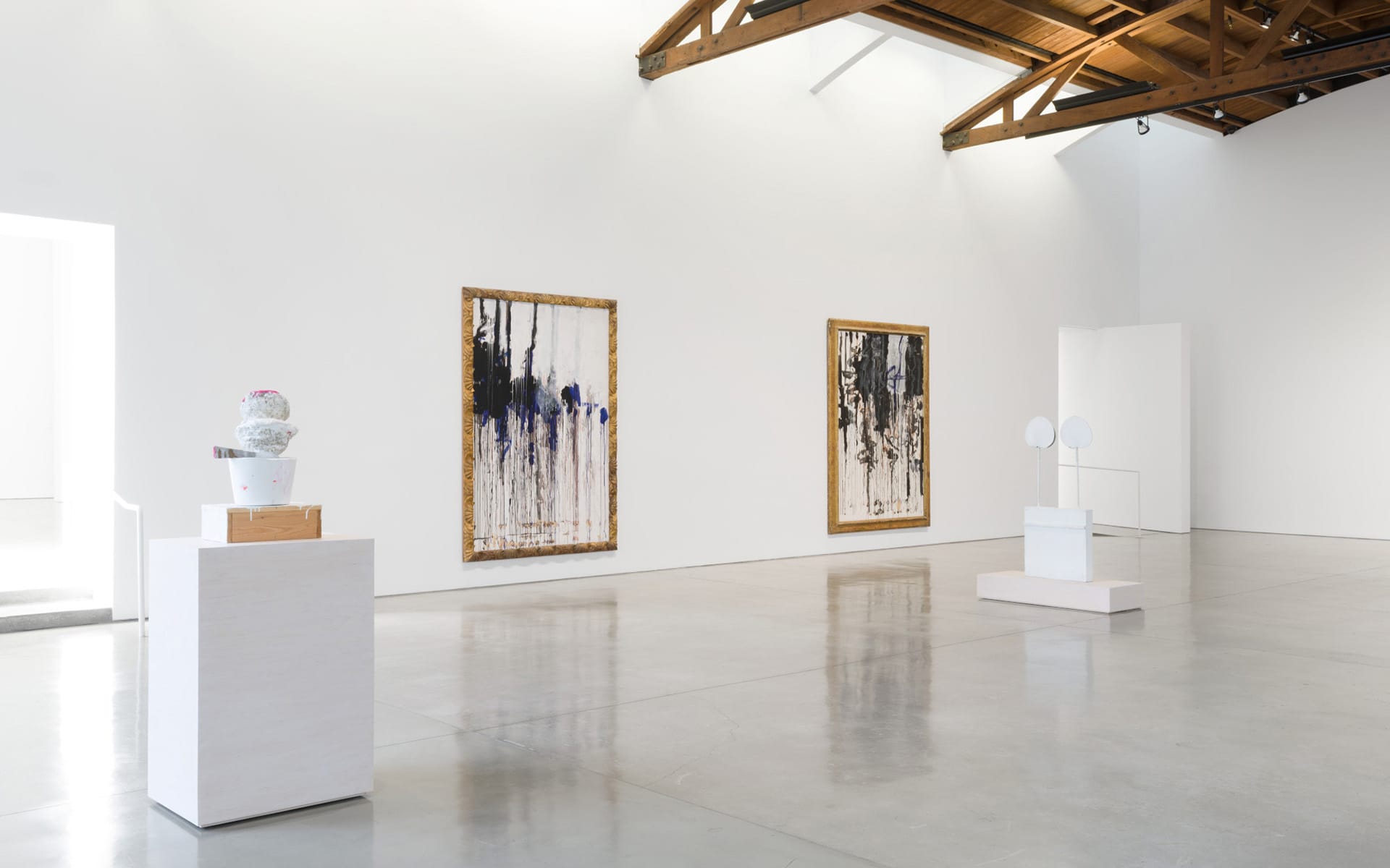 Art gallery interior with large abstract paintings on white walls, ceramic sculptures on pedestals, and natural light.