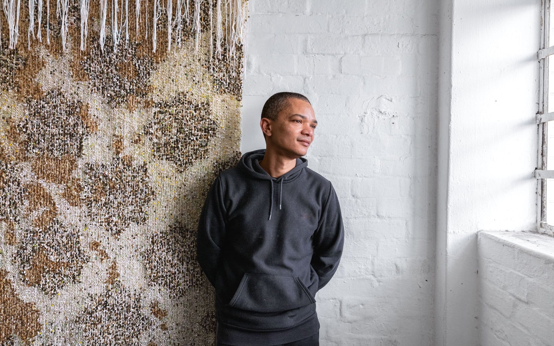 Person in a black hoodie leaning against a textured wall, looking out a window with soft lighting in a minimalist room.