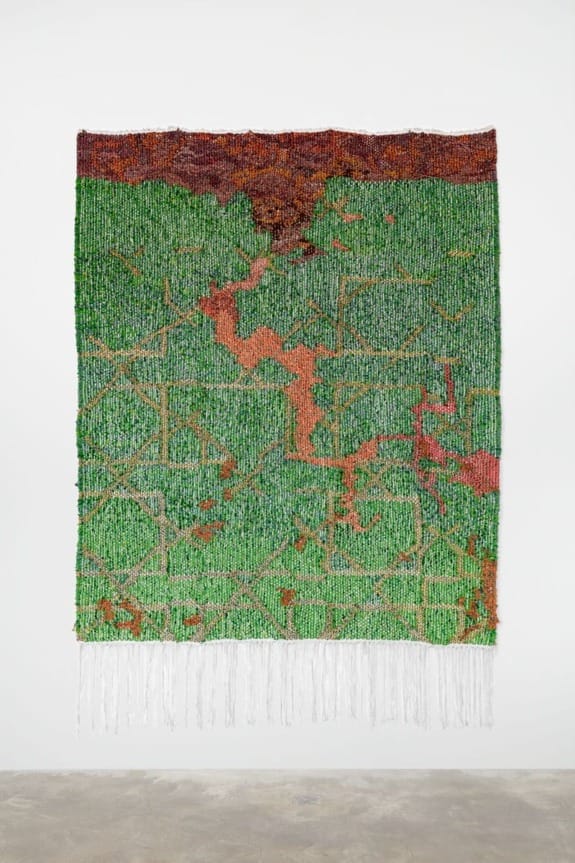 Abstract tapestry with green and orange patterns hanging on a white wall, featuring intricate textures and hanging threads below.