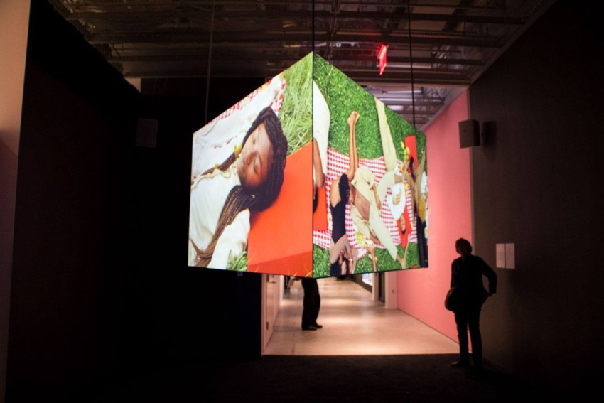 Art installation with suspended projectors displaying an outdoor picnic scene, observed by silhouettes in a dimly lit gallery.