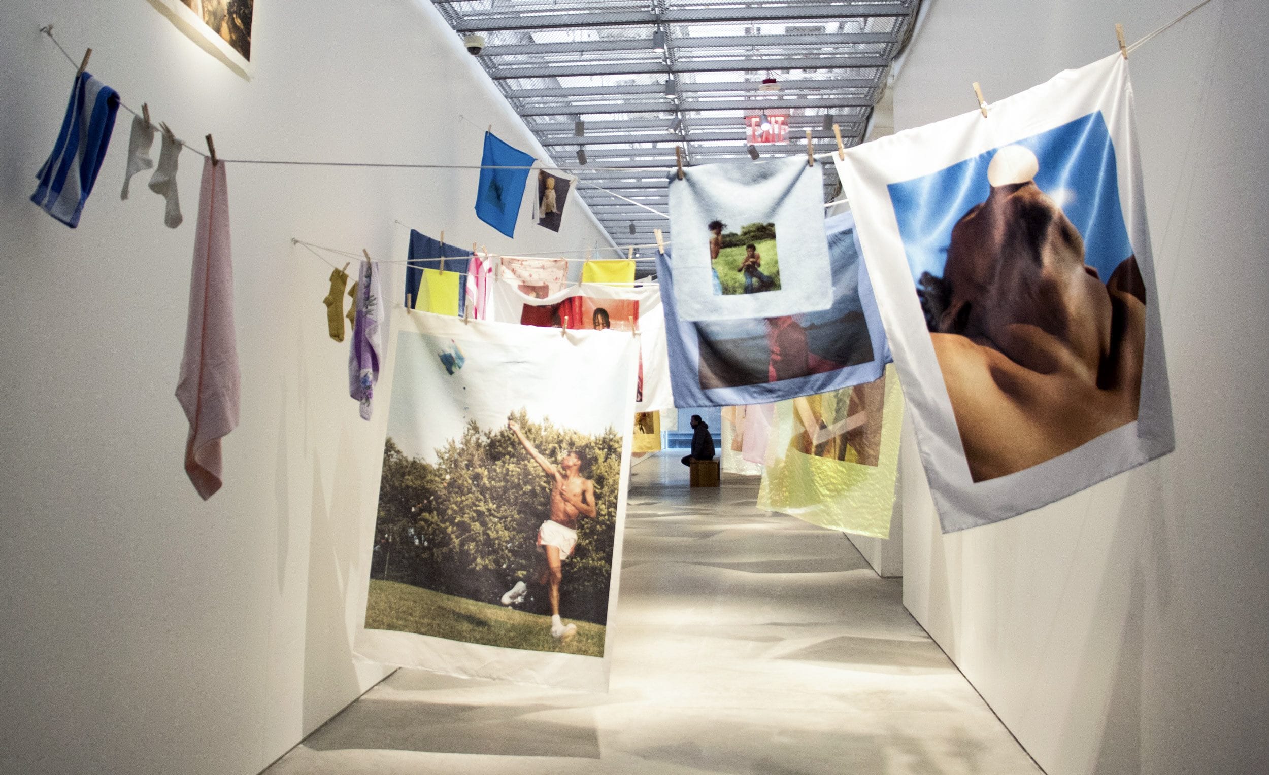 Art installation with large, colorful photographs hanging on clotheslines in a gallery hallway with a person in the distance.