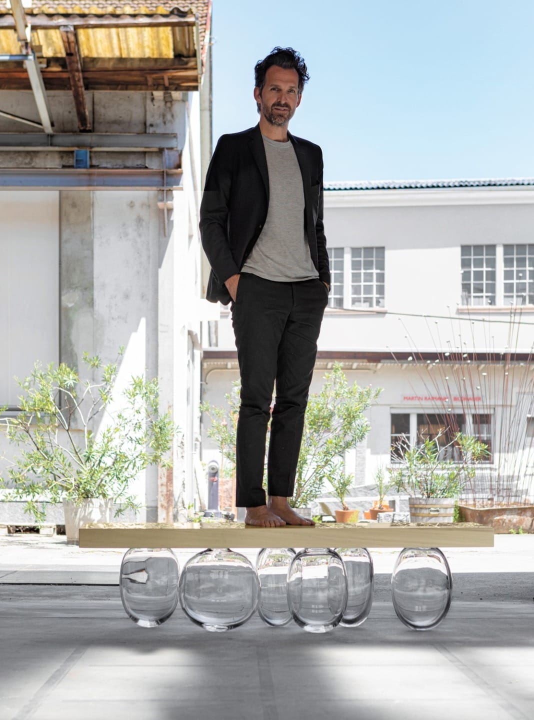Person standing on a wooden plank supported by clear glass vases in an industrial outdoor setting.