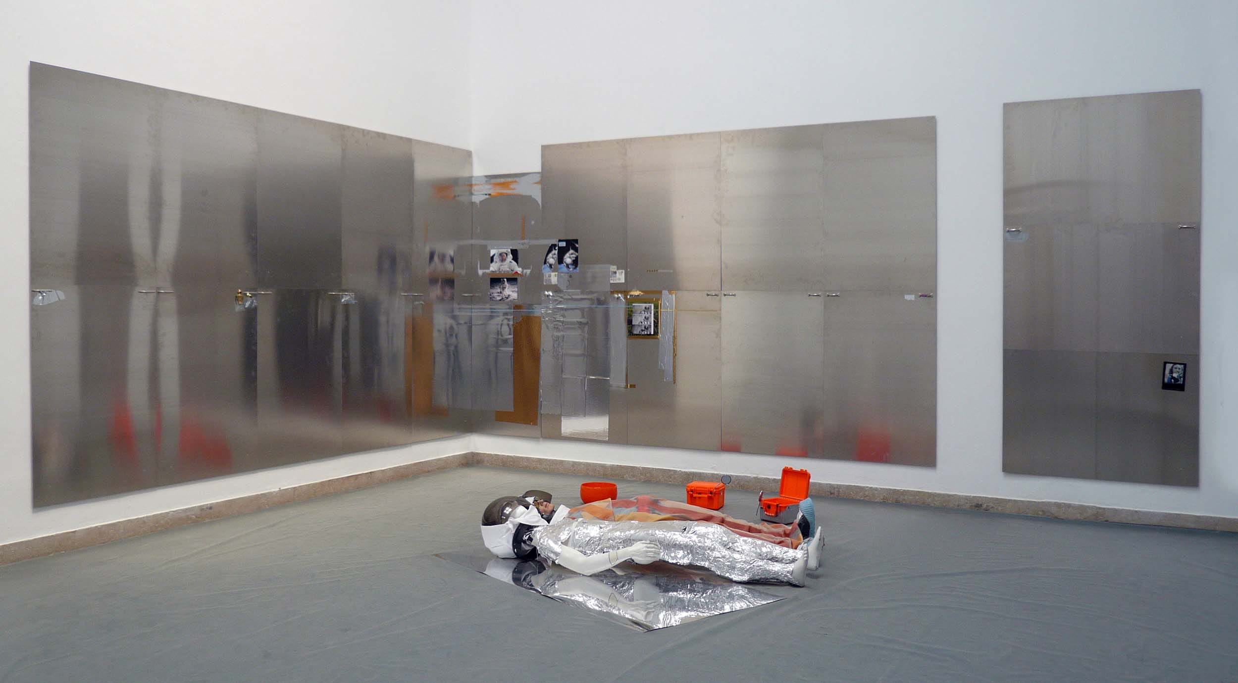 Modern art installation with reflective panels, floor objects resembling astronaut wrapped in shiny material, and orange buckets.