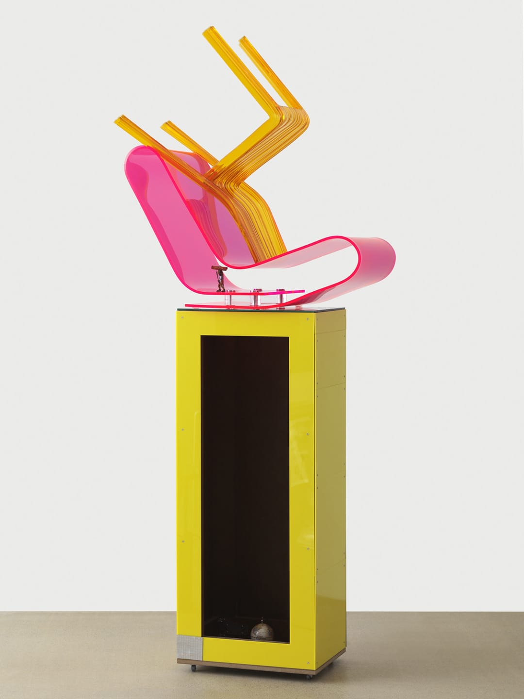Modern abstract sculpture with bright pink and yellow geometric shapes on a tall yellow pedestal against a plain background.