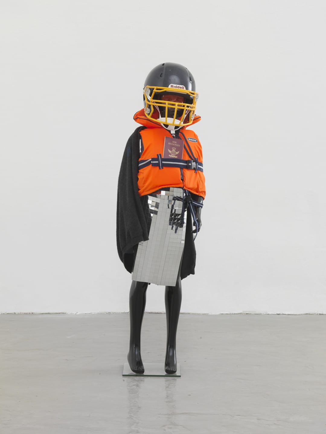 A mannequin wears a sports helmet, orange life jacket, black garment, and holds a reflective object in a minimalist room.