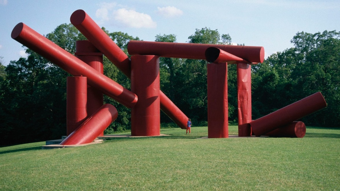 Large red abstract sculpture with cylindrical shapes on a grassy field under a blue sky, person standing nearby for scale.