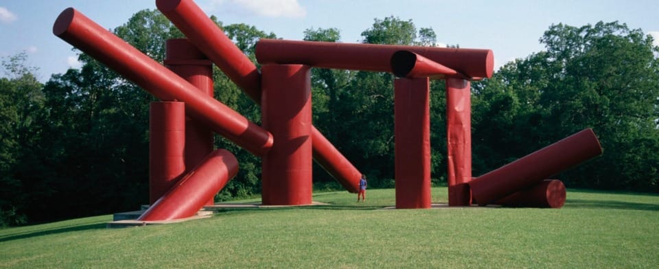 Large red abstract sculpture with cylindrical shapes on a grassy field under a blue sky, person standing nearby for scale.
