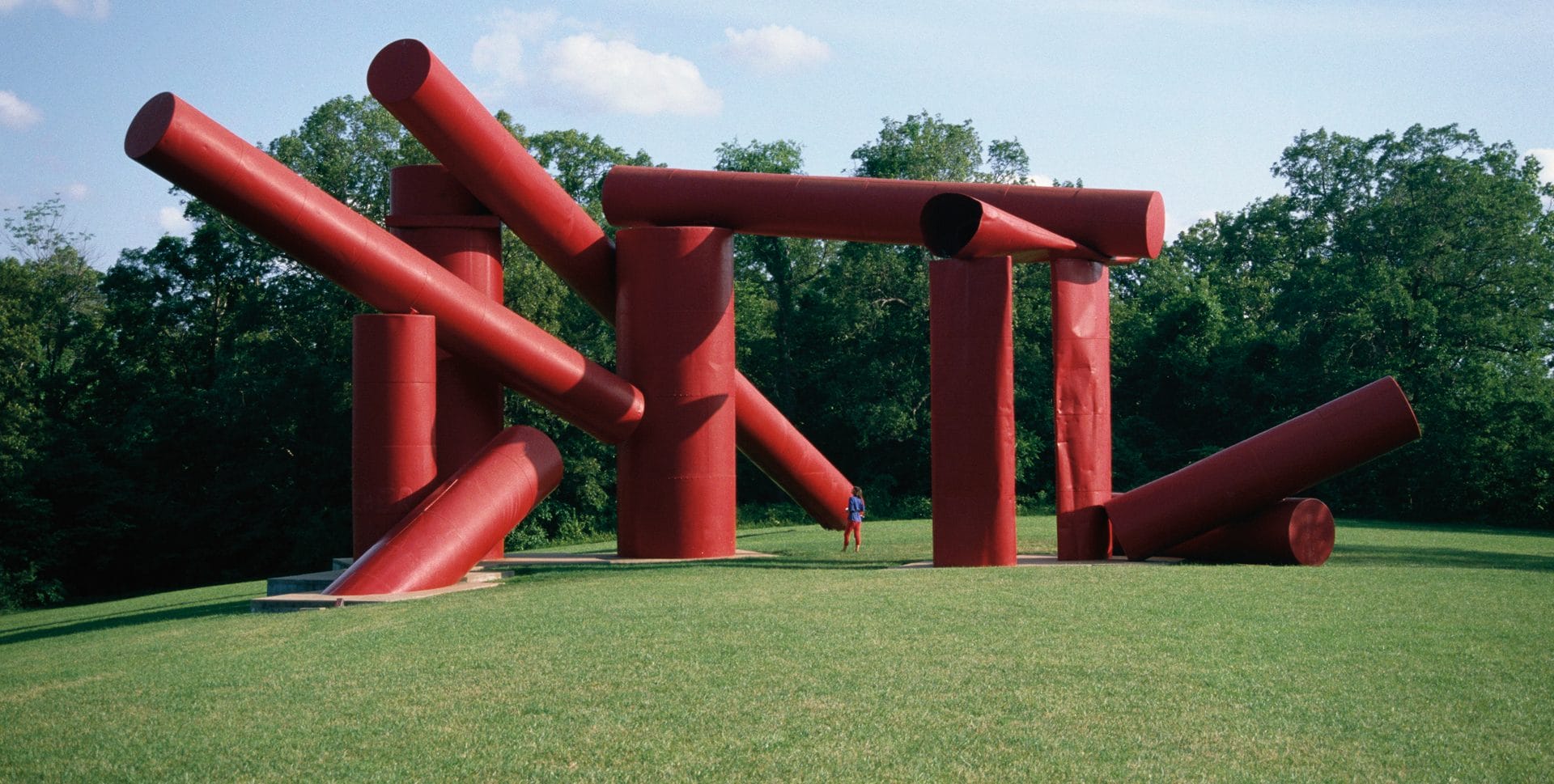 Large red abstract sculpture with cylindrical shapes on a grassy field under a blue sky, person standing nearby for scale.