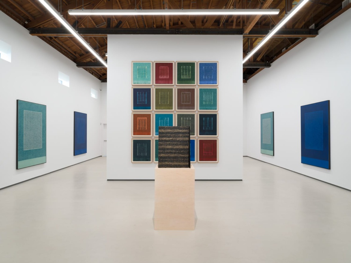 Contemporary art gallery interior with colorful abstract paintings on white walls and a central sculpture on a pedestal.