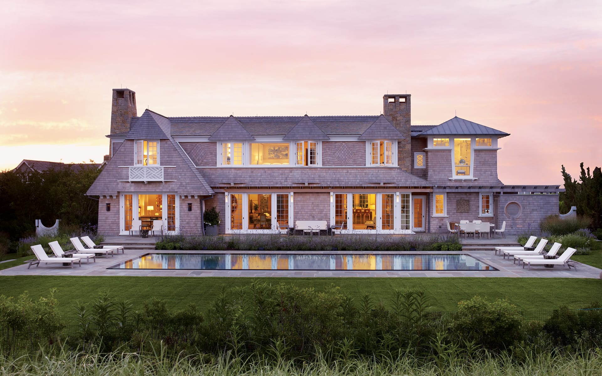 Elegant two-story house with large windows and a pool in the foreground at sunset.