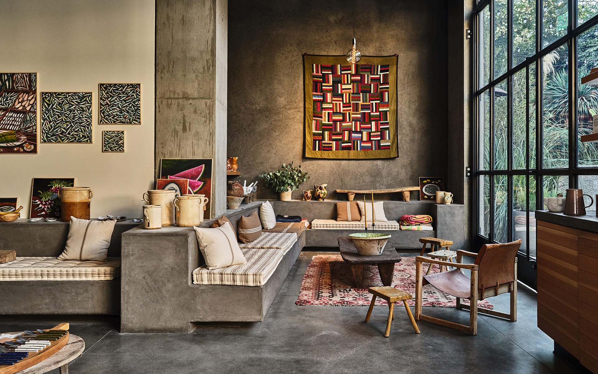 Modern living room with concrete seating, colorful wall art, and large windows. Decor includes plants and wooden furniture.