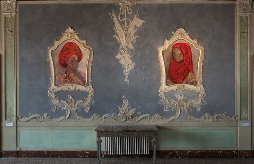 Vintage-style room with ornate frames on a gray wall featuring two abstract portraits in red attire above a decorative table.