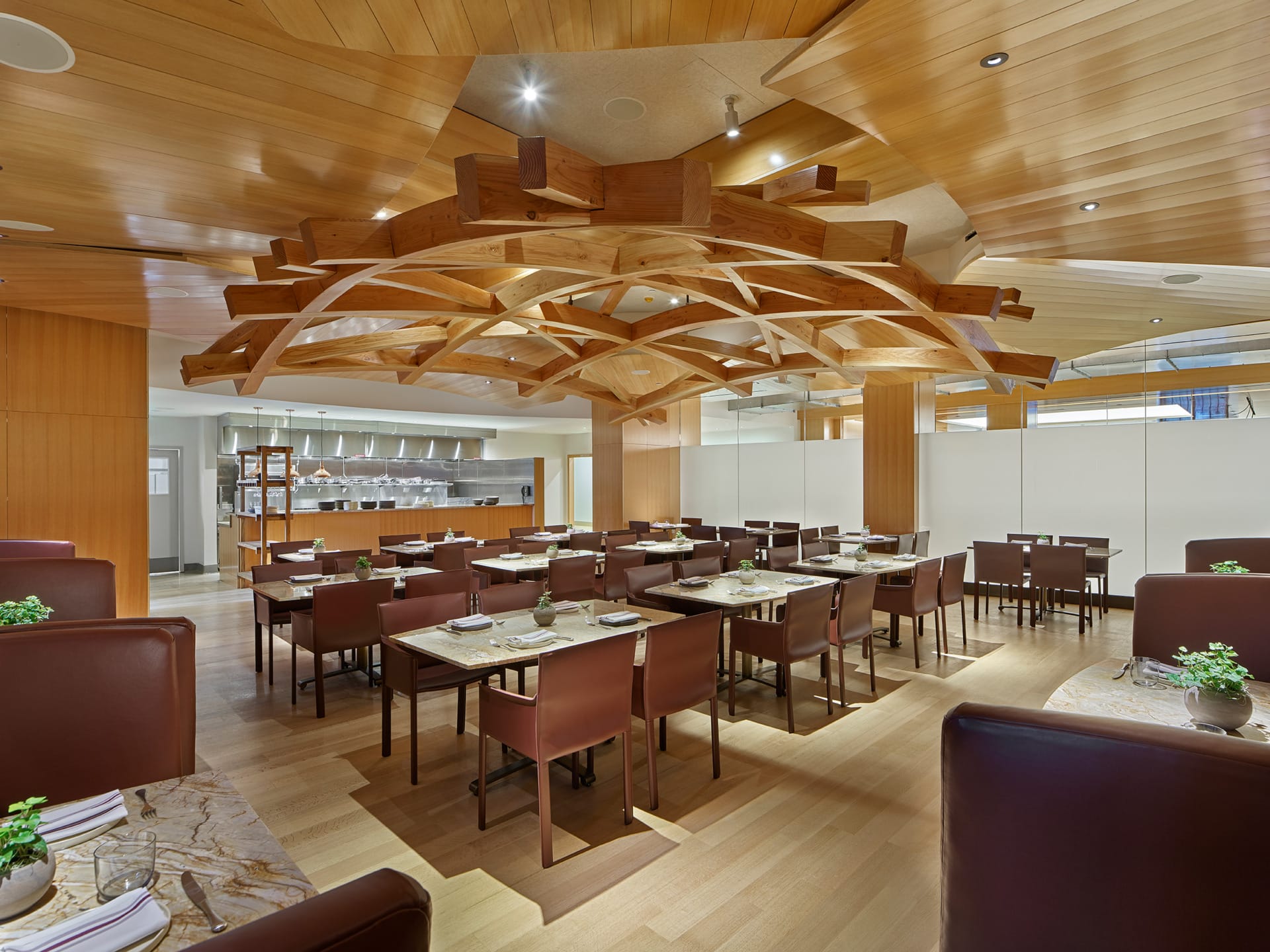 Modern restaurant interior with wooden ceiling, brown chairs, set tables, and warm lighting.