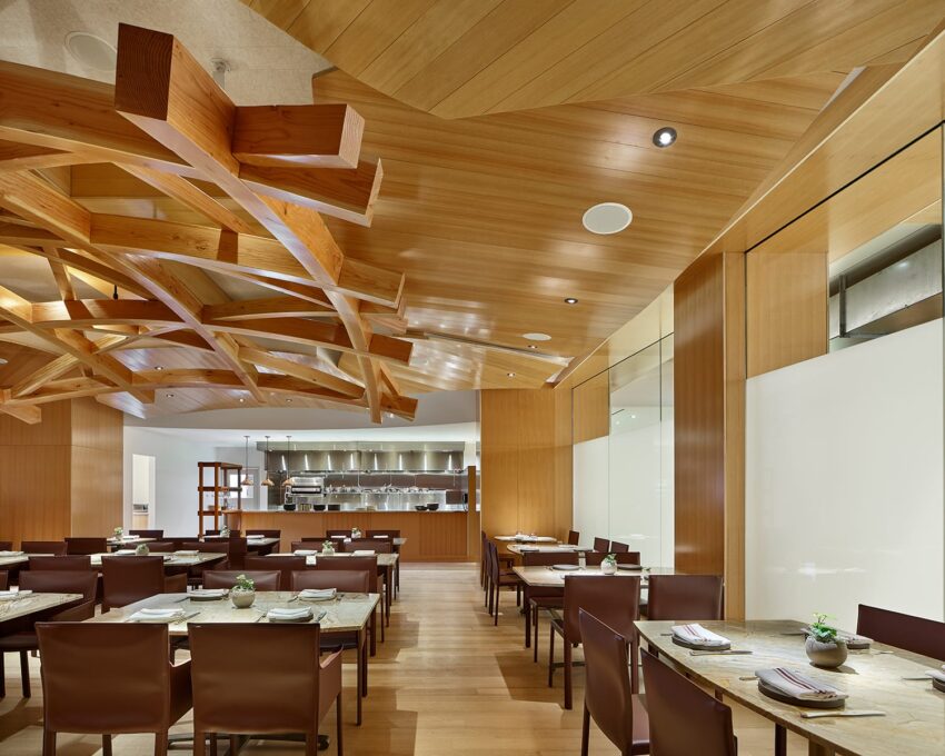 Modern restaurant interior with wooden ceiling, elegant tables, and open kitchen area in the background.