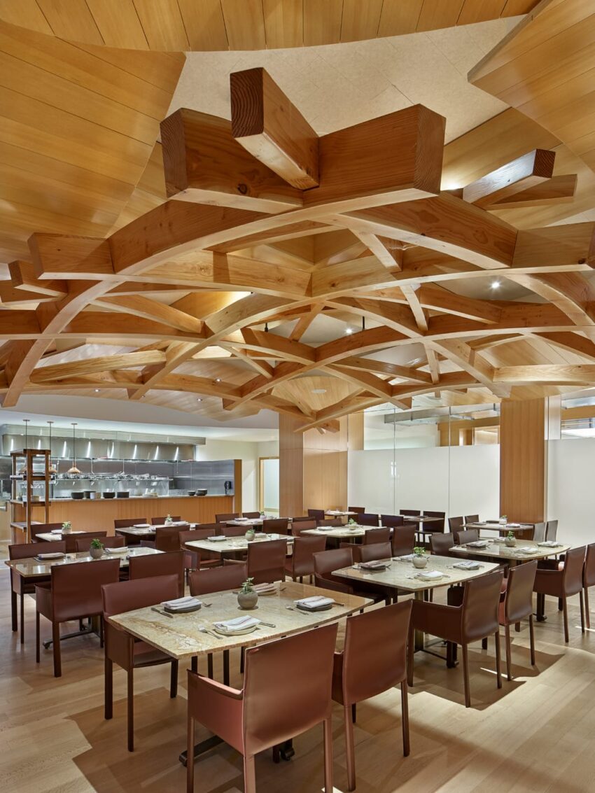 Modern restaurant interior with wooden ceiling design, tables set for dining, and open view of the kitchen in the background