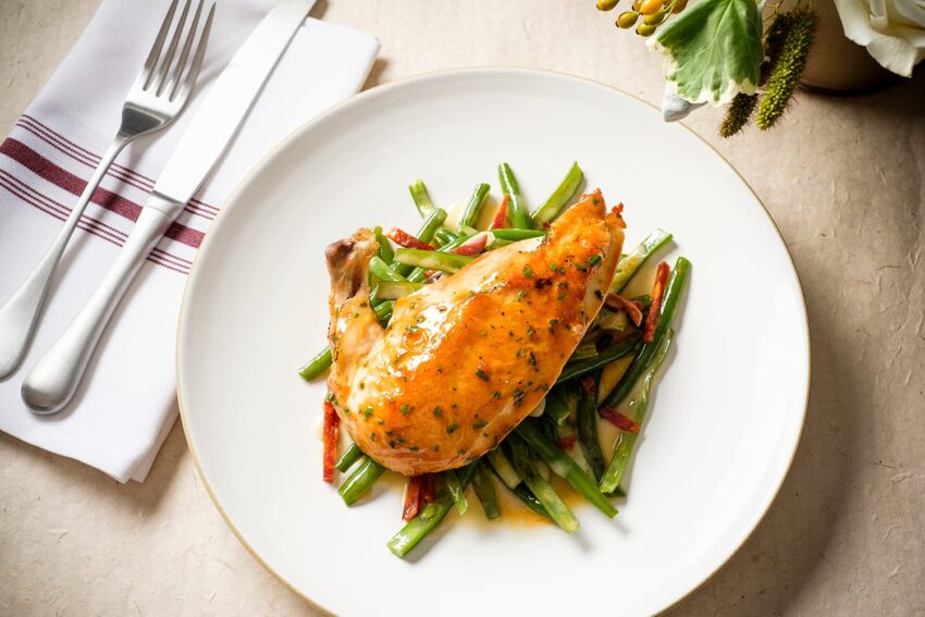 Grilled chicken leg on a bed of green beans and red peppers, served on a white plate with a fork and knife nearby.