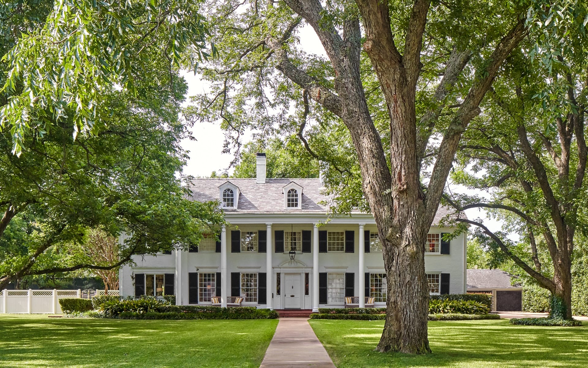 Historic colonial-style white house surrounded by lush green trees and a manicured lawn on a sunny day.
