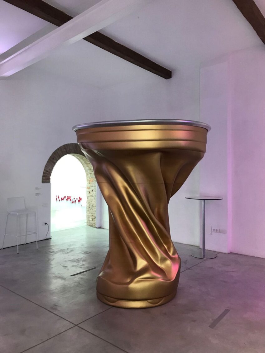 Large twisted golden can sculpture in a modern art gallery with white walls and an arched doorway in the background.