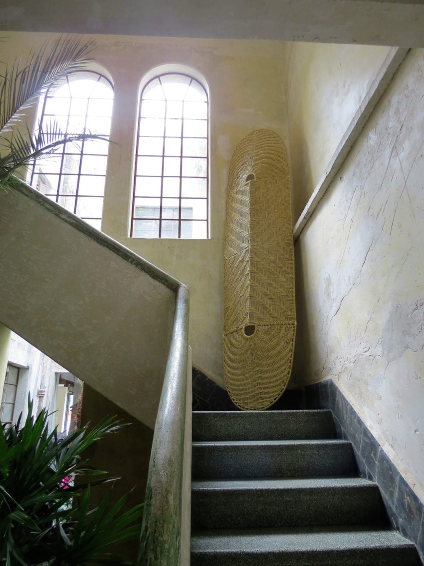 Staircase with textured walls and large windows, featuring a tall decorative woven piece leaning against the wall.