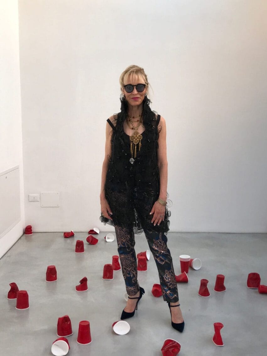 Person in a black outfit and sunglasses stands among scattered red cups on a gray floor in a white room.