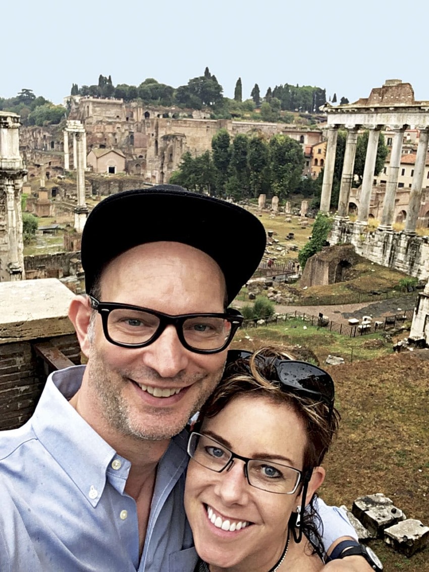 A couple smiling in front of ancient Roman ruins with columns and greenery in the background.