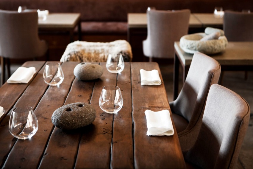 Rustic dining setup with wooden table, modern chairs, glasses, stone centerpieces, and neatly folded napkins in a cozy restaurant.