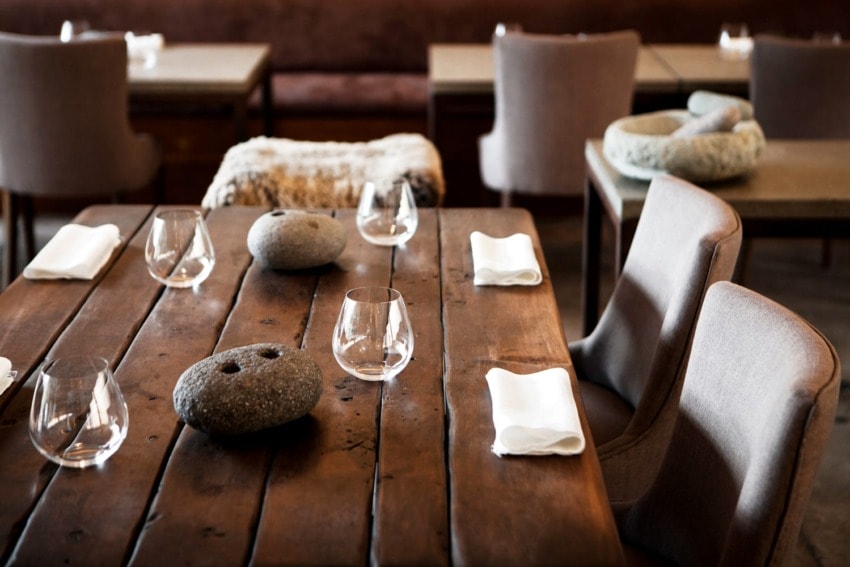 Rustic dining setup with wooden table, modern chairs, glasses, stone centerpieces, and neatly folded napkins in a cozy restaurant.