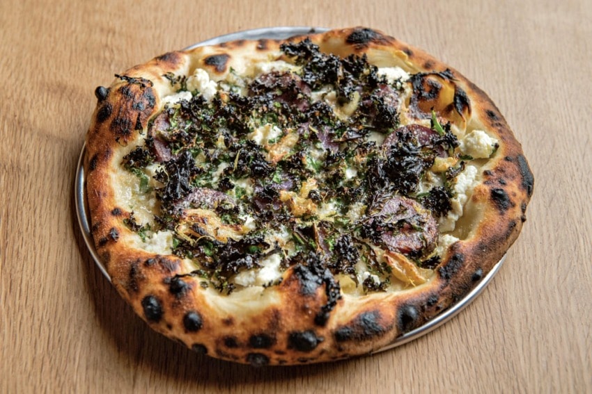 Wood-fired pizza topped with kale, ricotta cheese, caramelized onions, and purple potato slices on a metal tray.