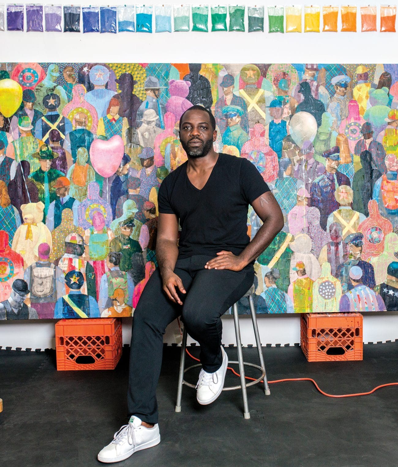 Artist sitting on a stool in front of a colorful abstract painting, with a row of paint swatches above.