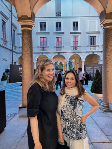 Two women smiling in a stylish outdoor courtyard with elegant arches and evening gathering in the background.