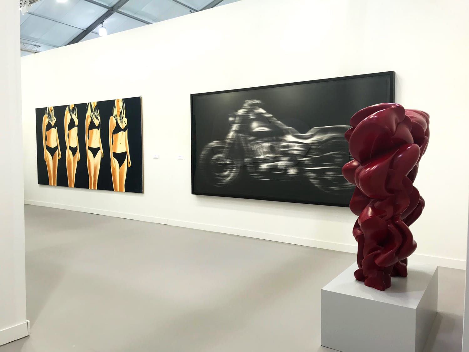Art gallery with a red abstract sculpture and paintings of a motorcycle and women in swimsuits on a white wall.