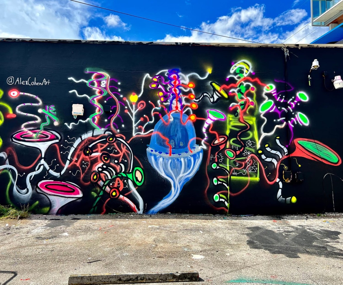Colorful street art mural with abstract shapes and a central blue jellyfish against a black wall under a blue sky.