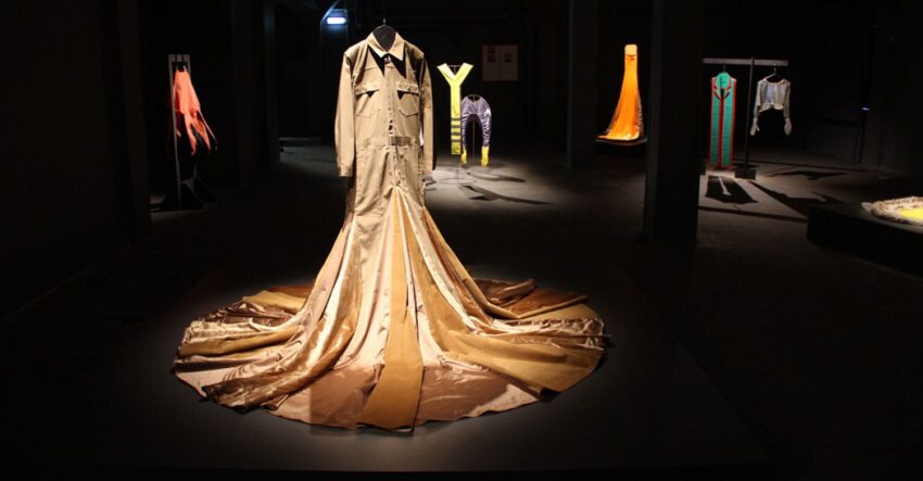 Exhibit of evening gowns with dramatic lighting, featuring a prominent golden gown as the centerpiece in a dark room.